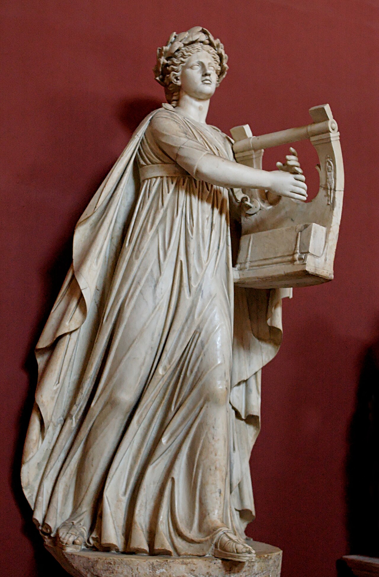 A full standing marble Apollo Musagetes, crowned with foliage and holding a kithara, against a deep red museum wall.