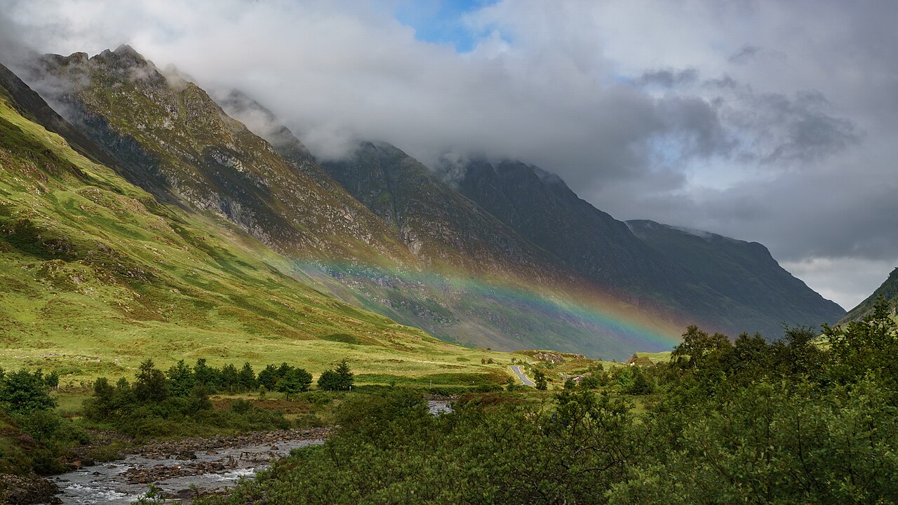 A rainbow crosses green slopes beneath cloud-covered mountains in Glen Coe.