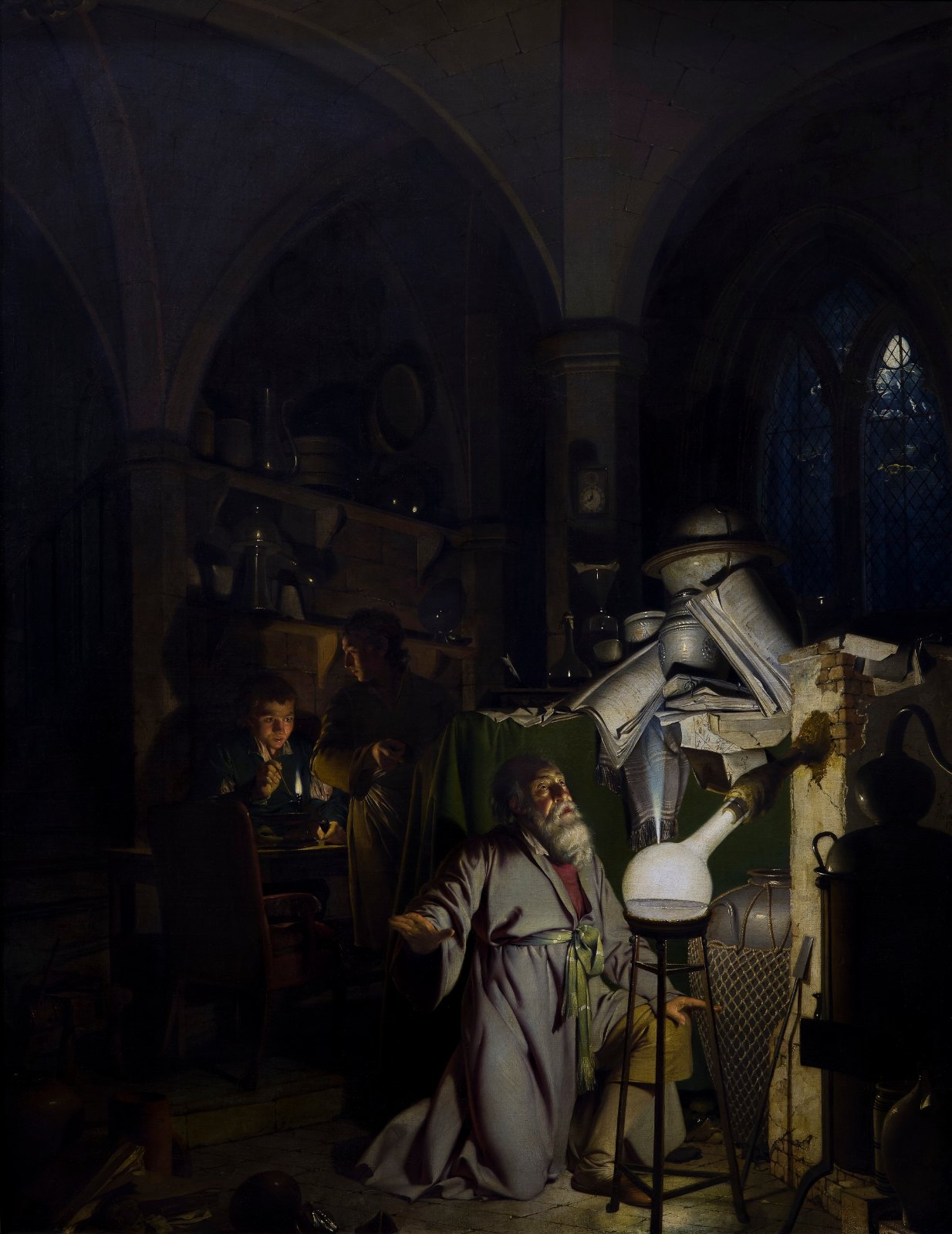A dark Gothic laboratory at night: an old alchemist kneels in prayer beside a glass flask that blazes with brilliant white light, while 2 assistants work at a table in the shadows behind him.