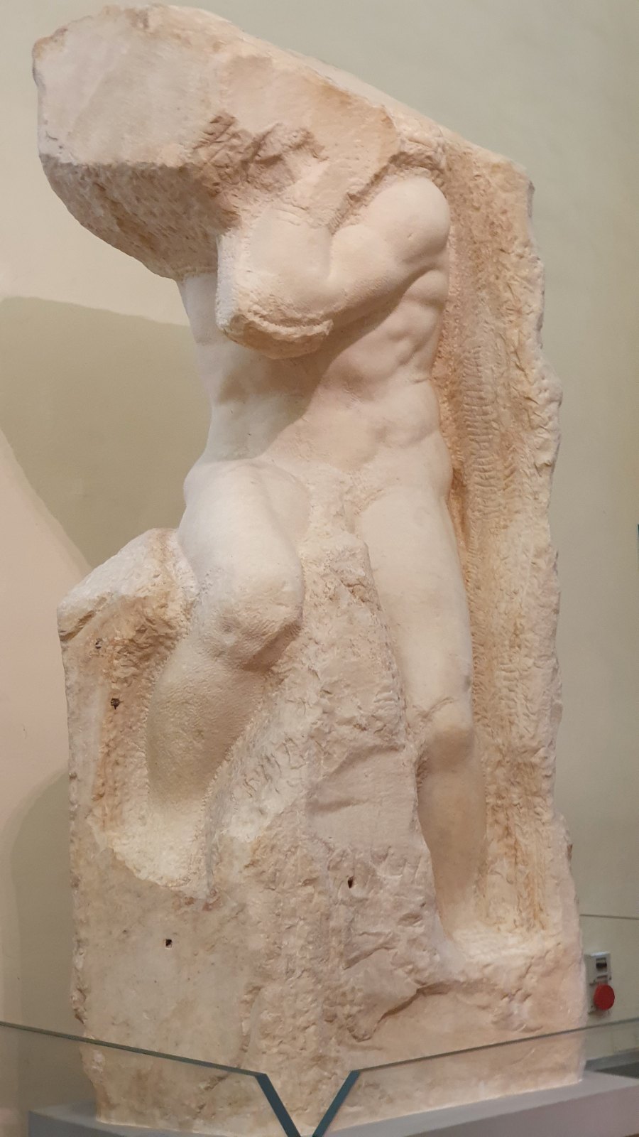 An unfinished marble figure: a muscular body bends under the weight of a mass of rough stone where his head should be, his arms raised to hold it, his legs only partly freed from the block.