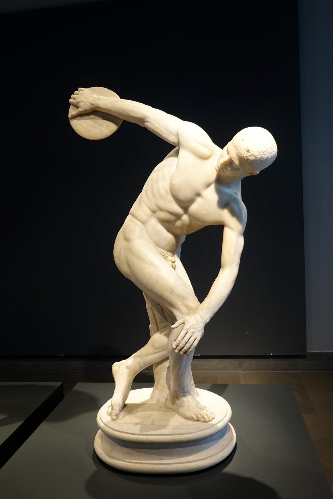 A white marble statue of a nude athlete twisted in the instant before releasing a discus, spot-lit against a dark background.