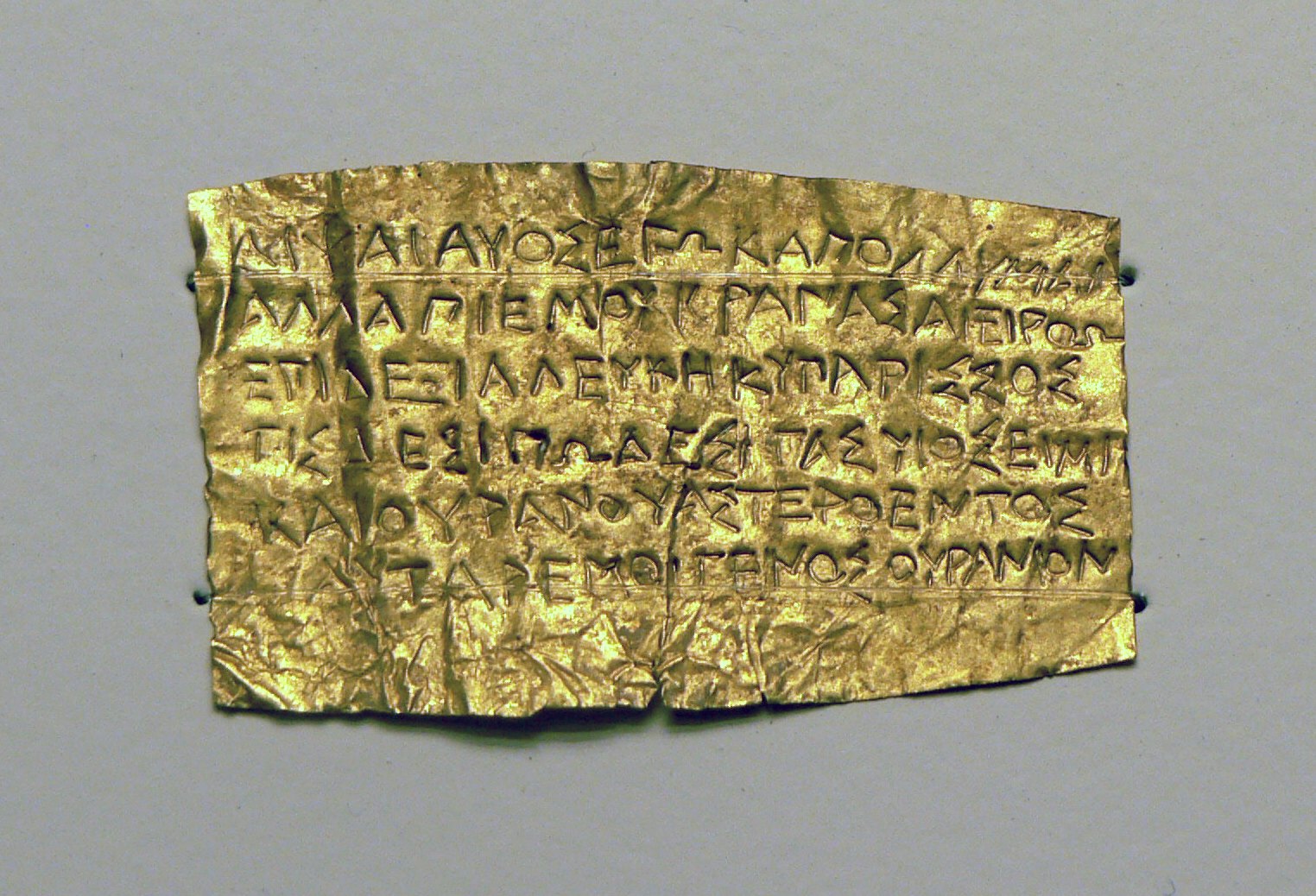 A small, thin sheet of gold leaf, crumpled at the edges, inscribed with several lines of Greek capital letters.