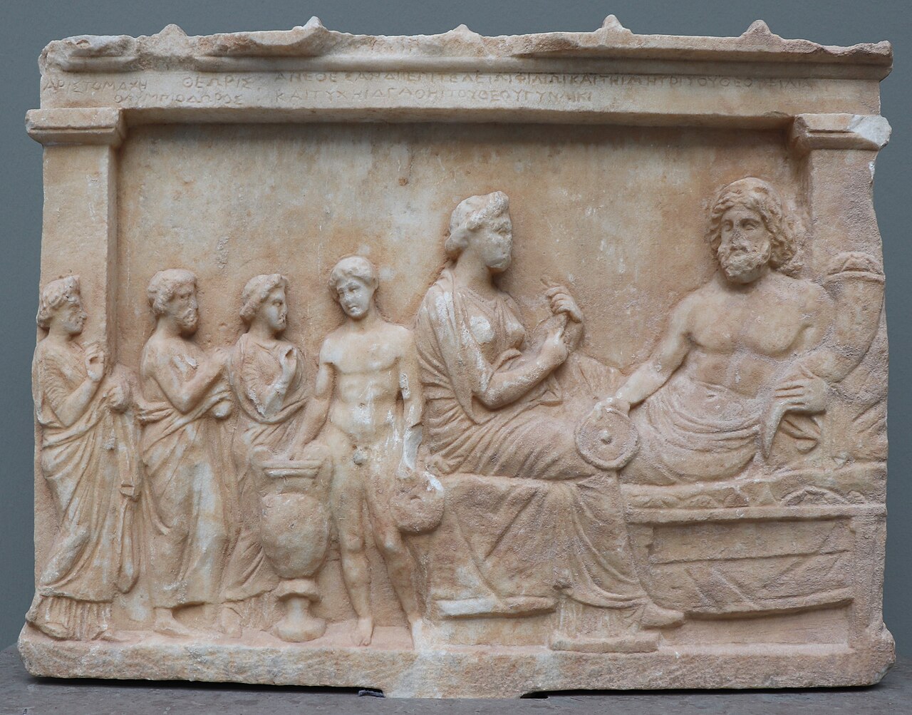 Marble votive relief: Zeus reclines on a banquet couch holding a horn of plenty and an offering bowl, a goddess seated beside Him, while worshippers approach from the left