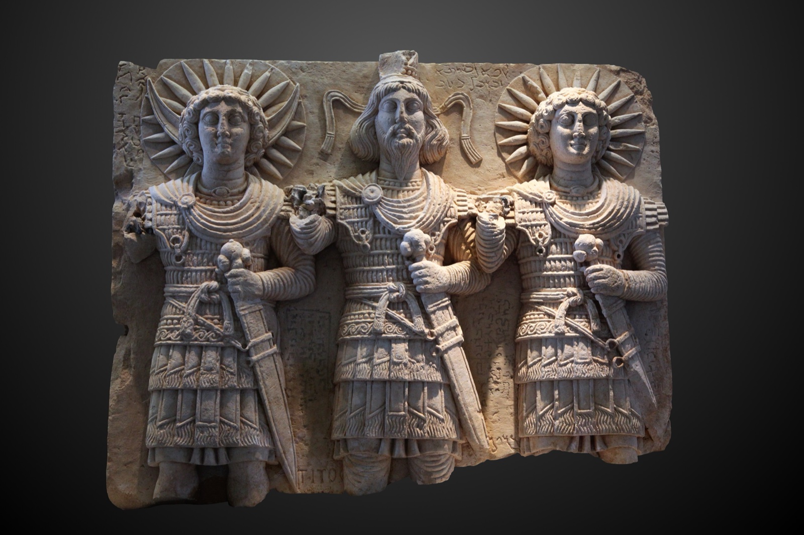 A limestone relief of three standing gods in armour and cloaks, the two outer figures with radiate halos and a crescent behind the shoulders of one.