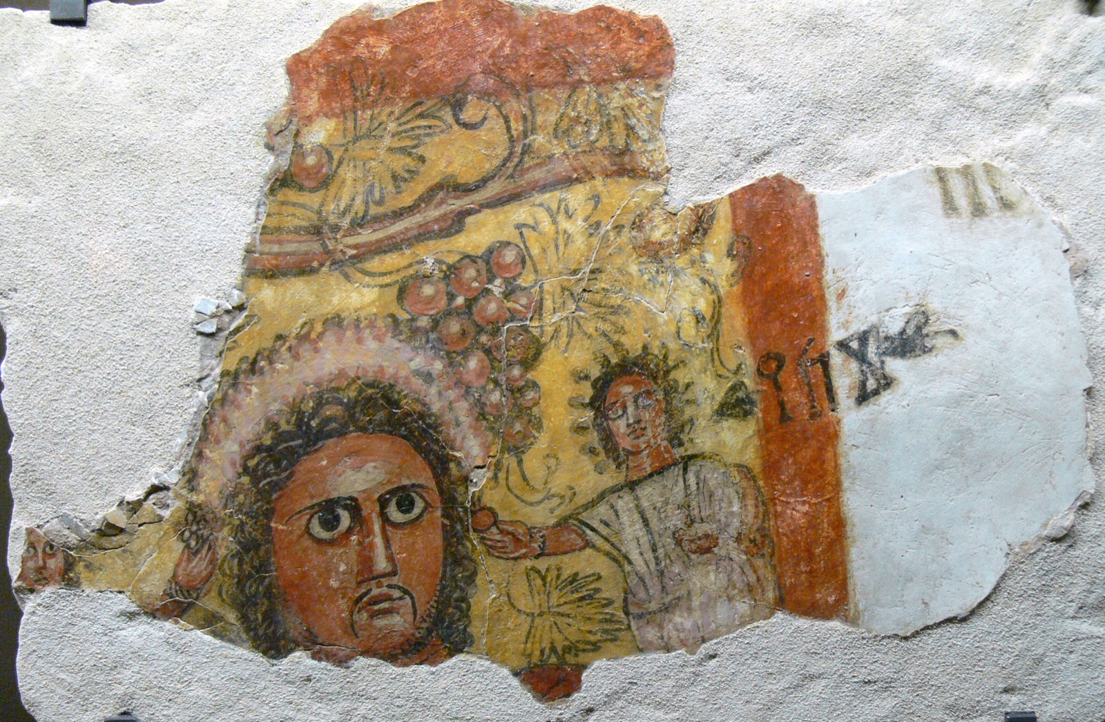 A fragment of painted wall plaster showing a man's face with large eyes and curling hair, a small second figure beside him and a short inscription in Ancient South Arabian letters.