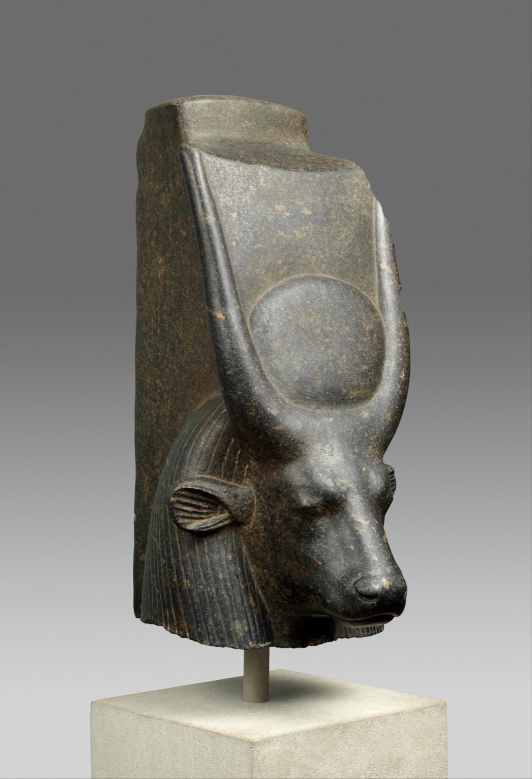 Dark stone head of a Goddess with the face of a cow and a sun disk between Her horns.