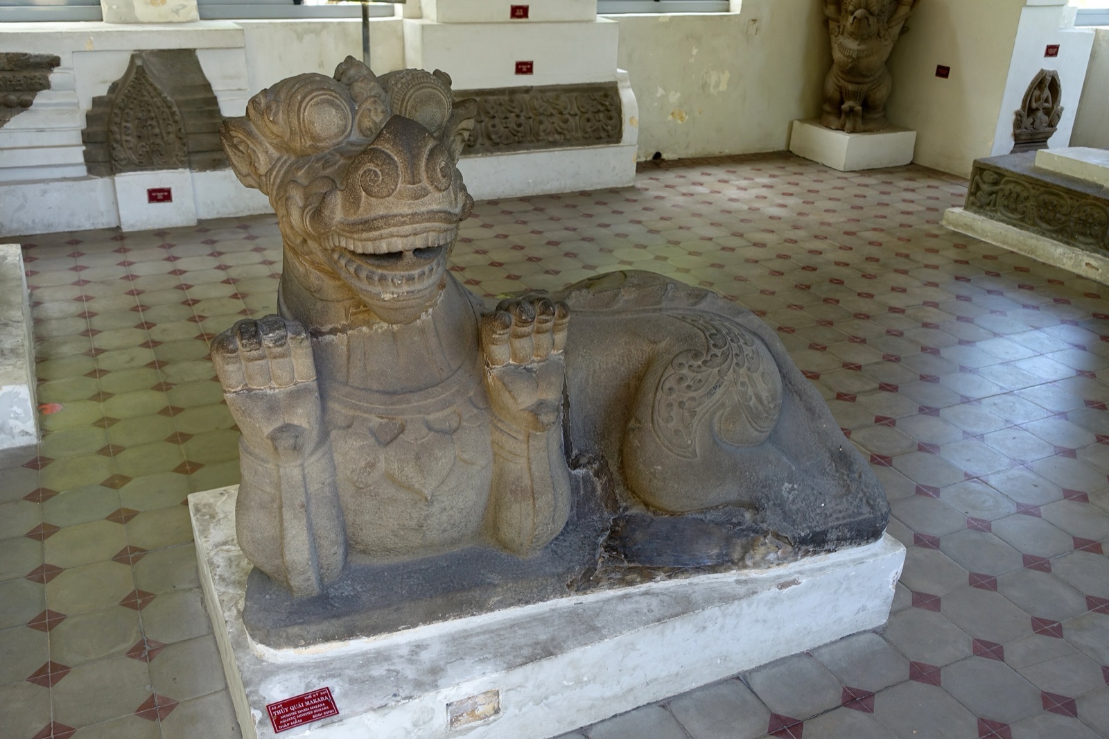 Weathered stone sculpture of a makara, a sea creature with a curled snout and open jaws, on a museum plinth.